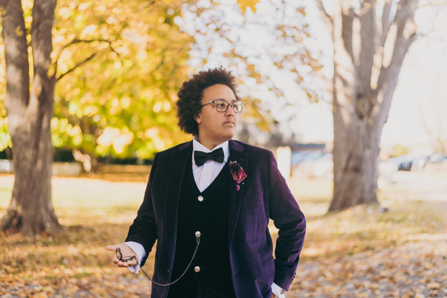 Groom in velvet navy suit with boutonniere poses outdoors among autumn trees.