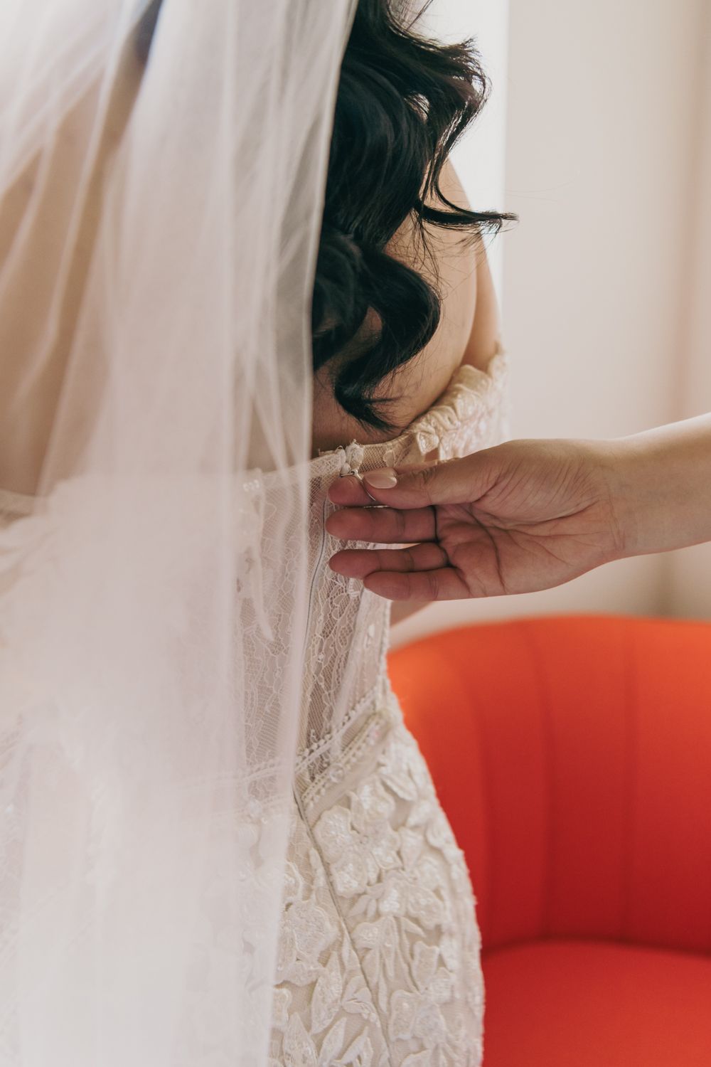 A helper fastens the back of a brides lace wedding dress while she wears a flowing veil.