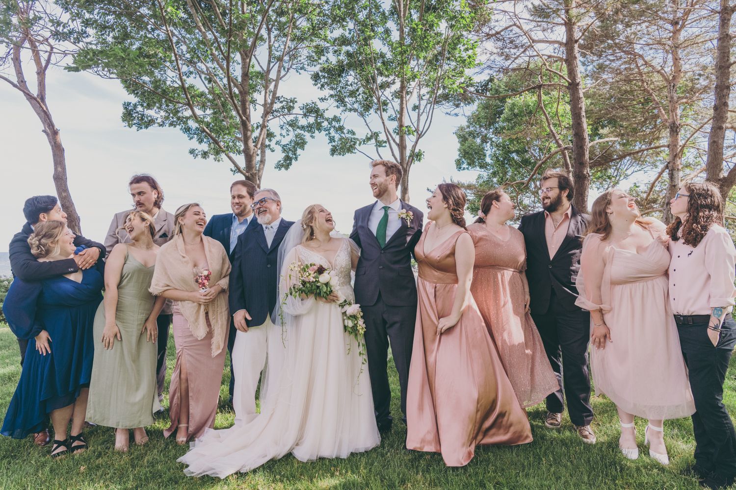 Wedding party in blush and navy gowns laughing with bride and groom under trees outdoors.
