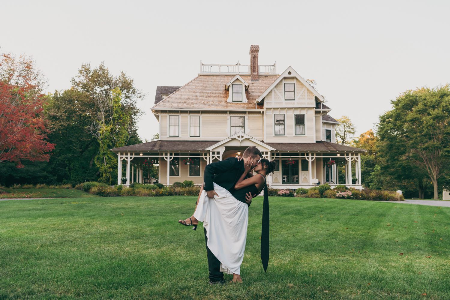 Couple sharing a romantic kiss in front of a charming Victorian house surrounded by lush green lawn.