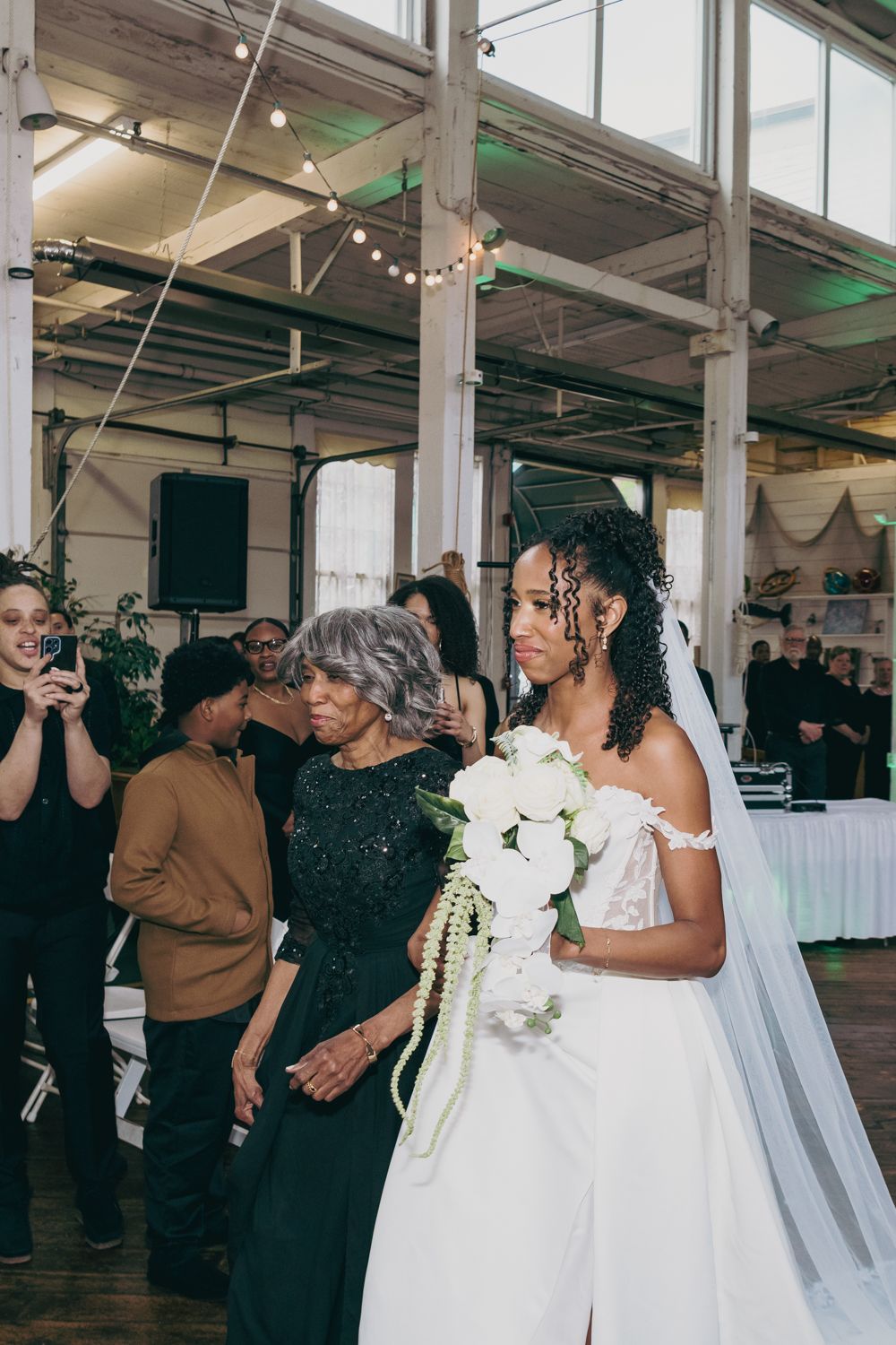 Bride in white ball gown with veil and bouquet walks down aisle with elderly woman in black lace dress.