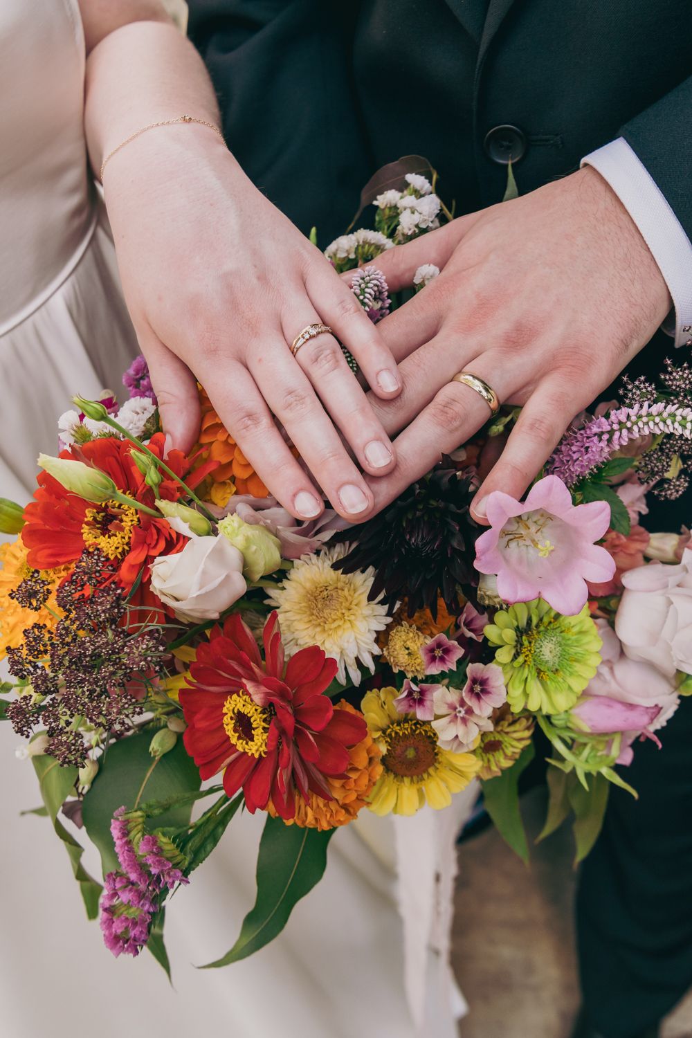Close-up of bride and groom's hands displaying wedding rings resting on a colorful bridal bouquet.
