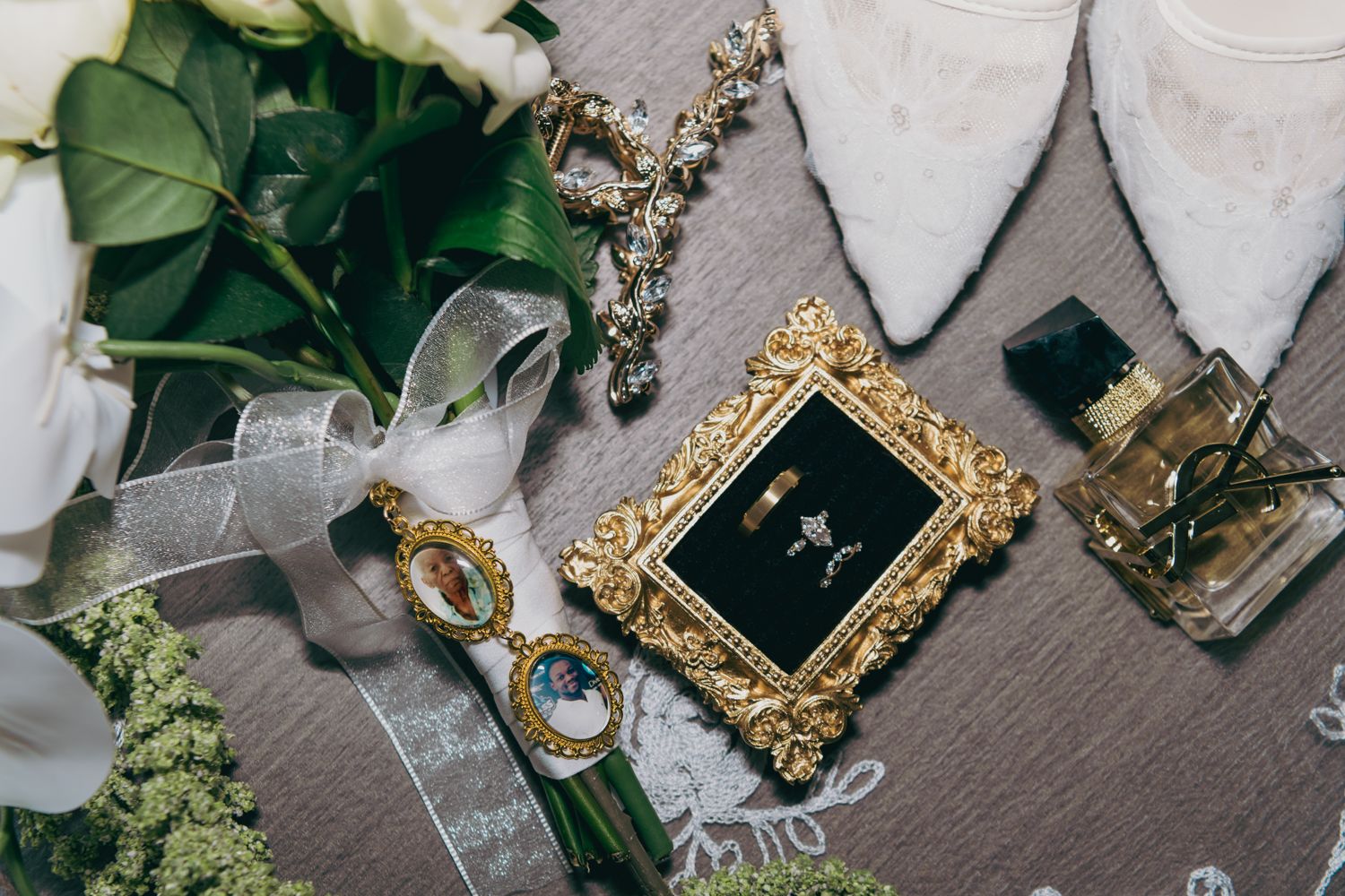 Elegant bridal flat lay featuring gold jewelry, perfume, wedding rings in ornate frame, white flowers, and lace ribbon.