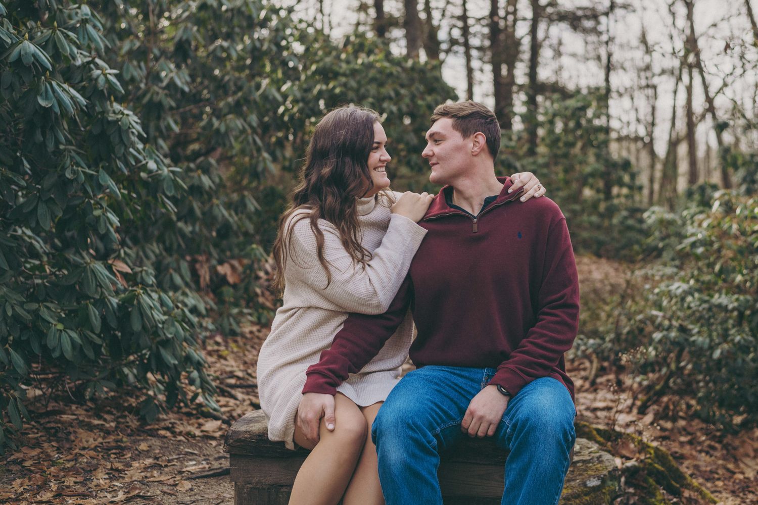A couple sits on a bench in a lush garden, smiling at each other in a romantic outdoor setting.