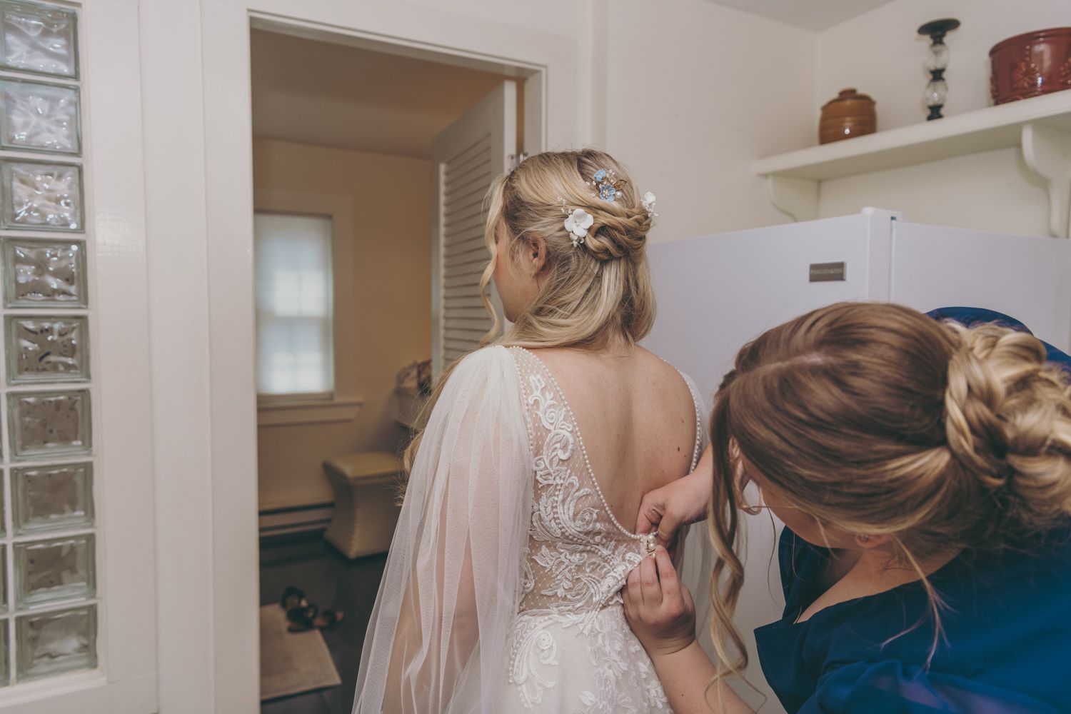 Bride in lace wedding dress having buttons fastened by bridesmaid in blue dress before wedding ceremony.