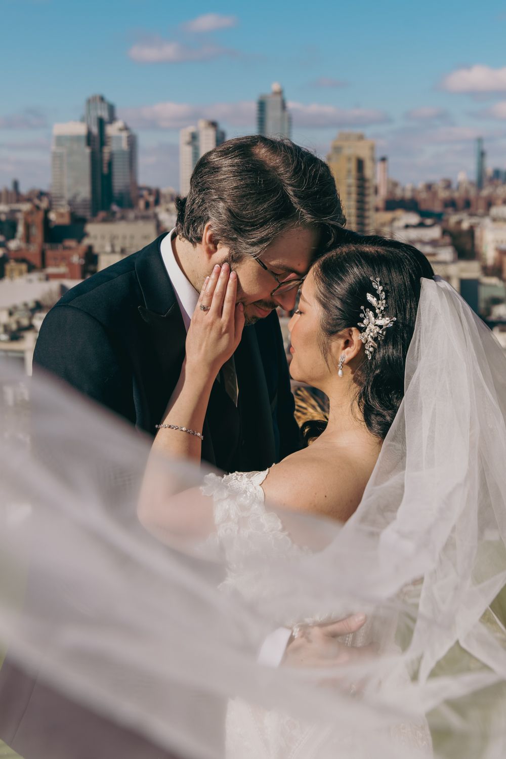 Bride and groom share a romantic rooftop moment with NYC skyline backdrop, veil billowing in the breeze.