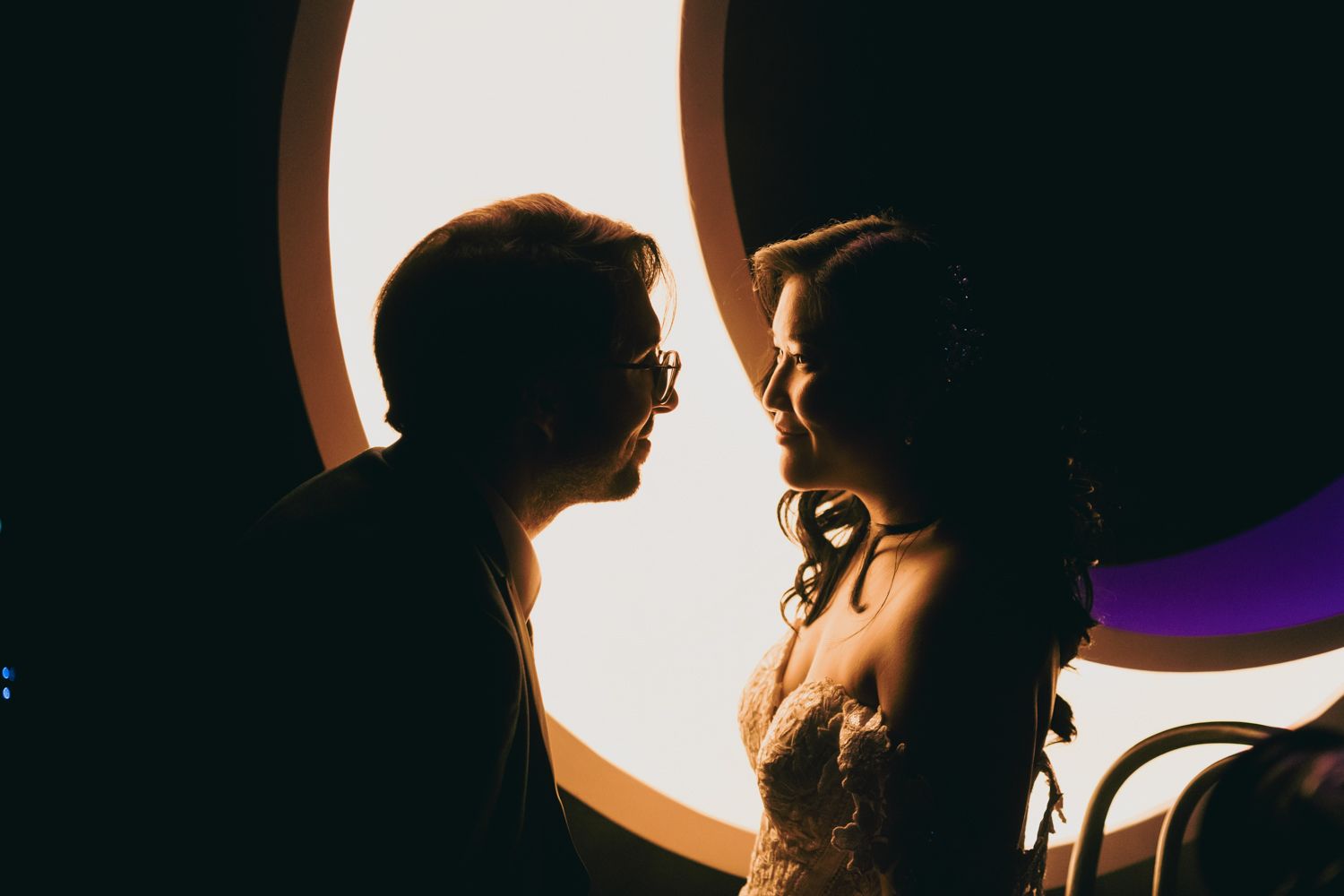 Romantic silhouette of bride and groom facing each other in front of a glowing circular light.