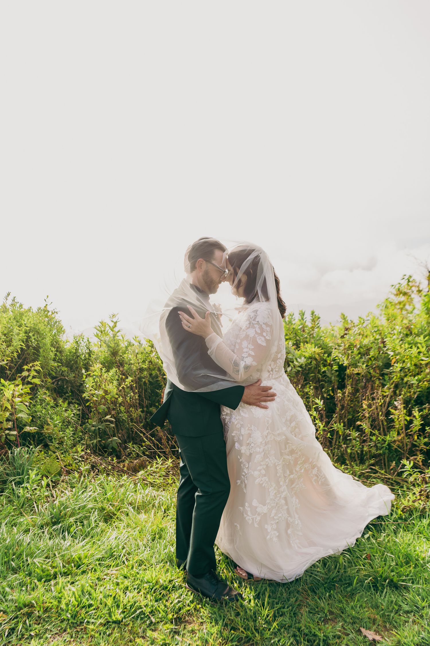 A bride and groom share a romantic embrace outdoors, surrounded by lush greenery, as her lace dress and veil flow in the breeze.