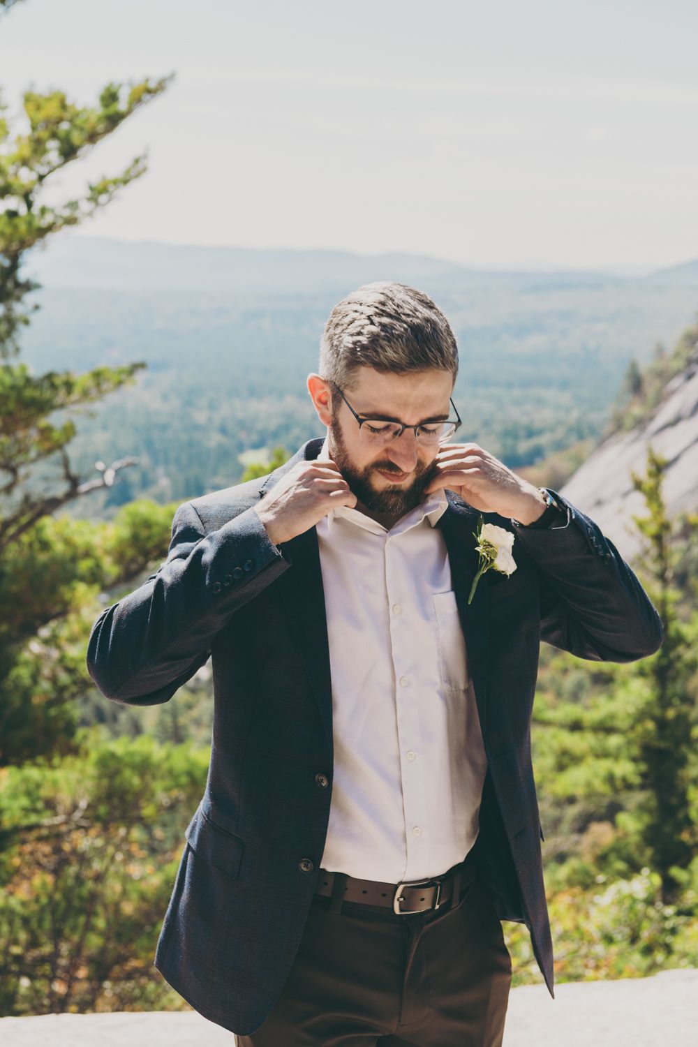 Handsome bearded groom in navy suit adjusting collar with mountain forest backdrop on wedding day.