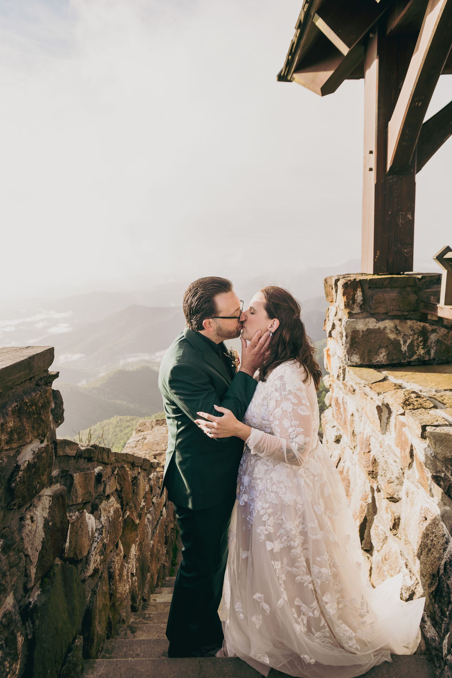 Newlyweds share a romantic kiss on a stone balcony with breathtaking mountain views in the background.