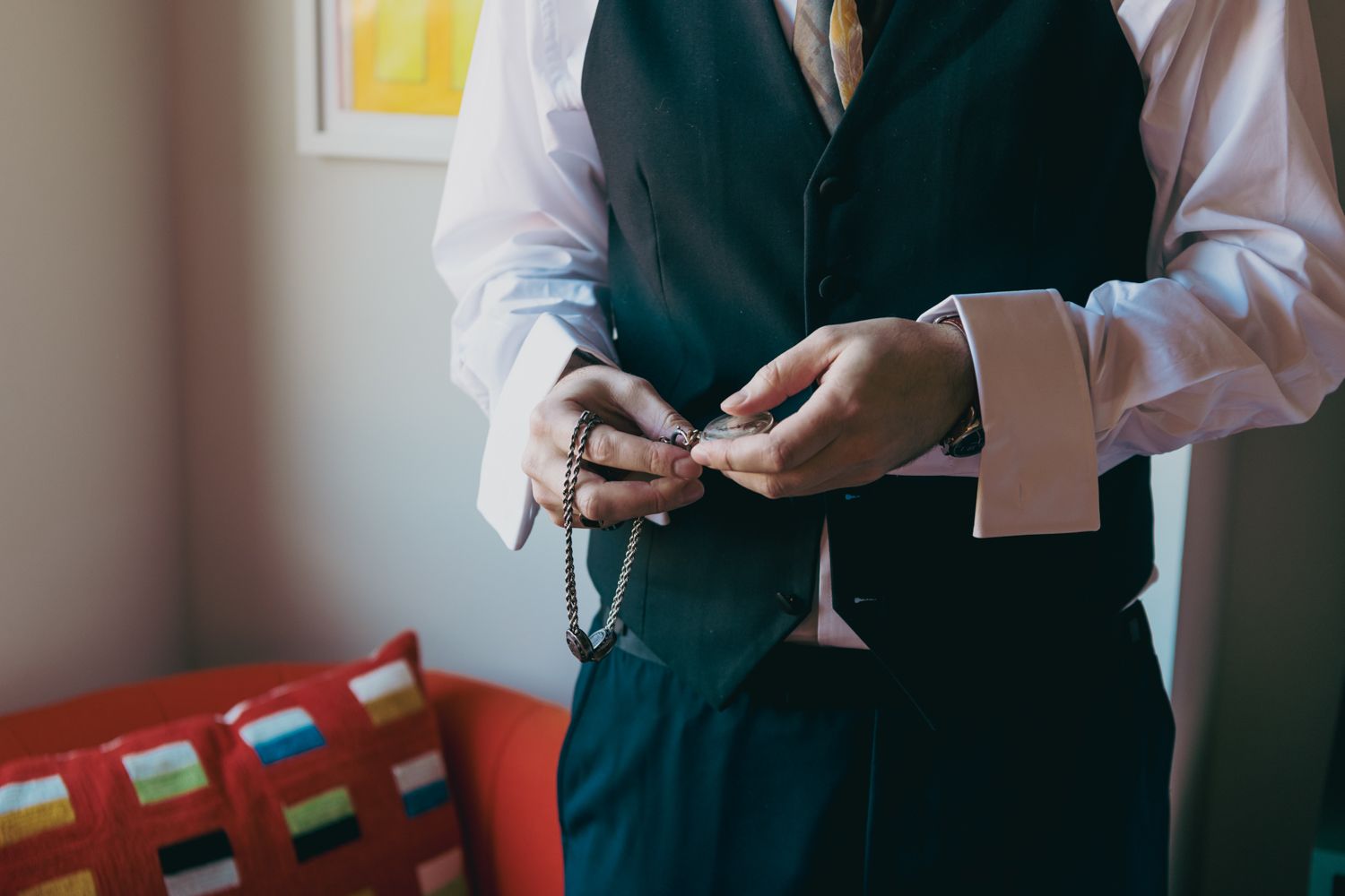 A groom in a navy vest and yellow tie fastens cufflinks while preparing for his wedding ceremony.