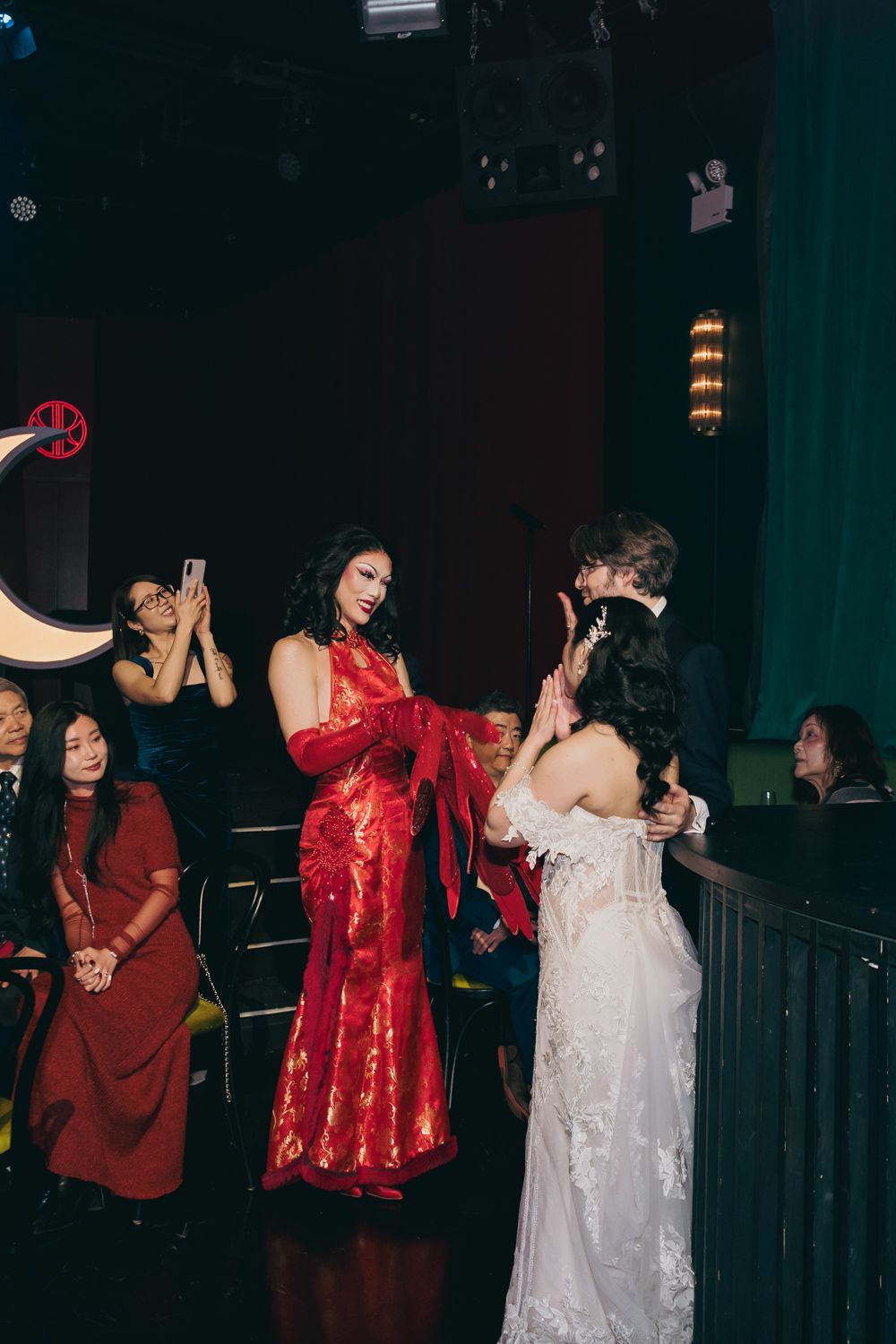 Woman in red gown interacting with bride in white lace dress at wedding.