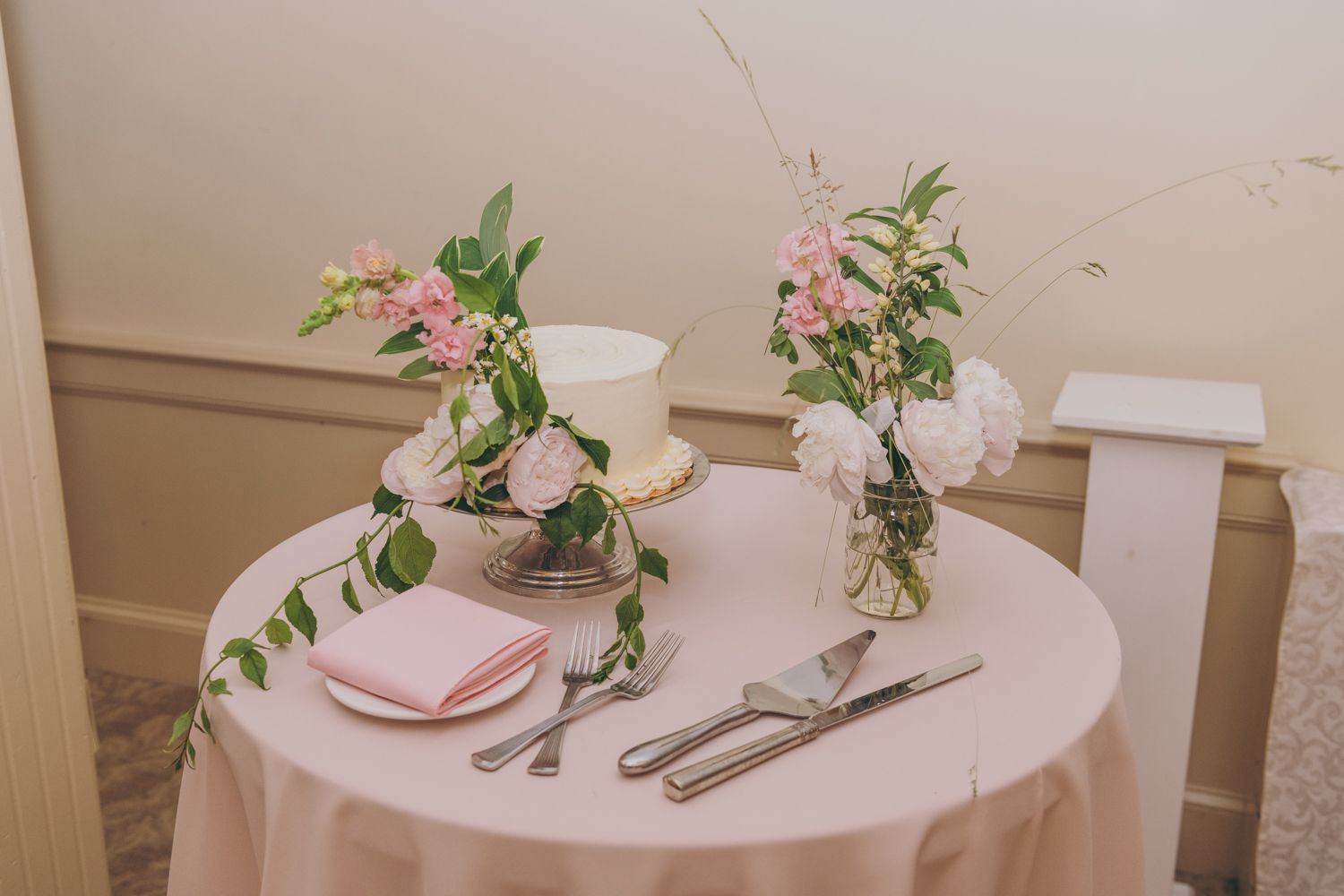 Elegant wedding cake table with pink roses, greenery, and silver serving set on blush tablecloth.