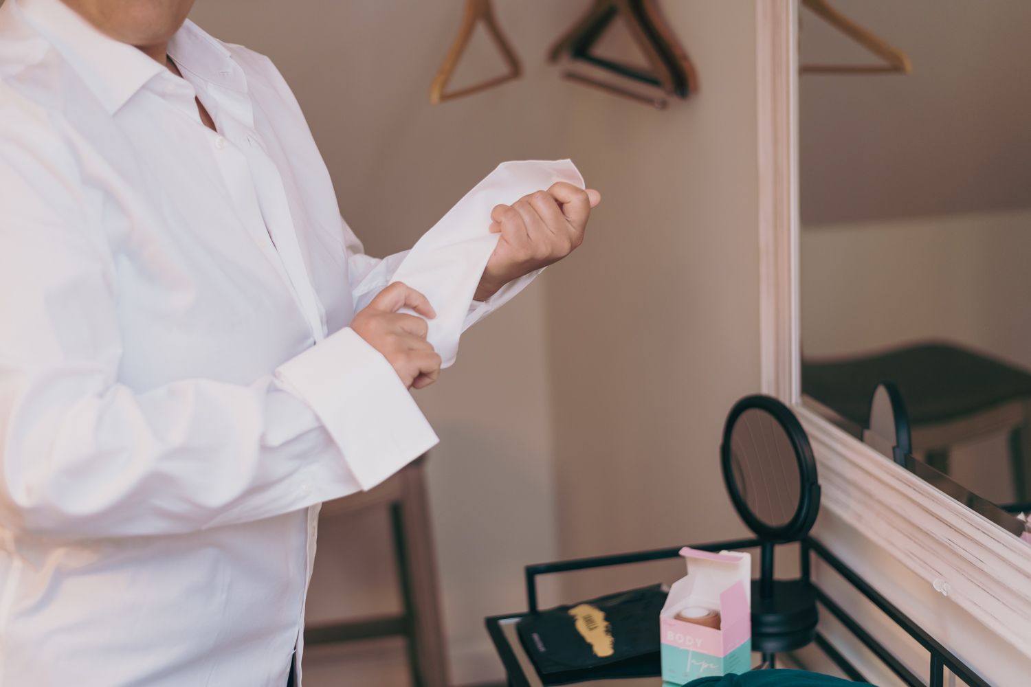 Man in white dress shirt buttoning cuffs while getting ready, with accessories on a vanity tray nearby.