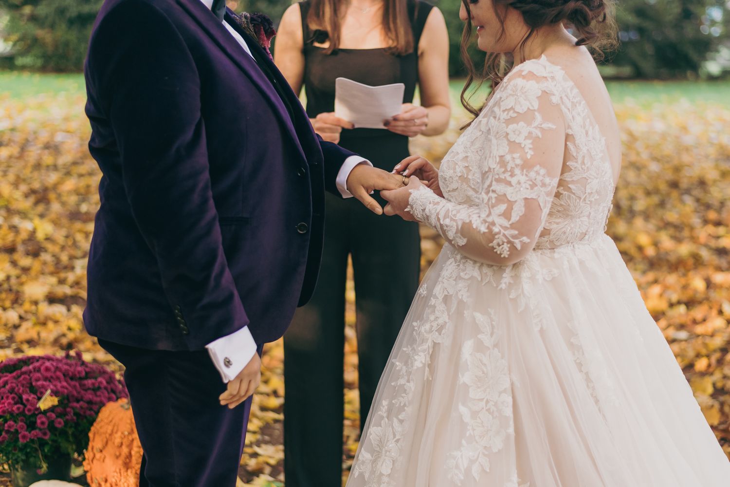 Couple exchanging rings during an intimate outdoor fall wedding ceremony officiated by a woman in black.