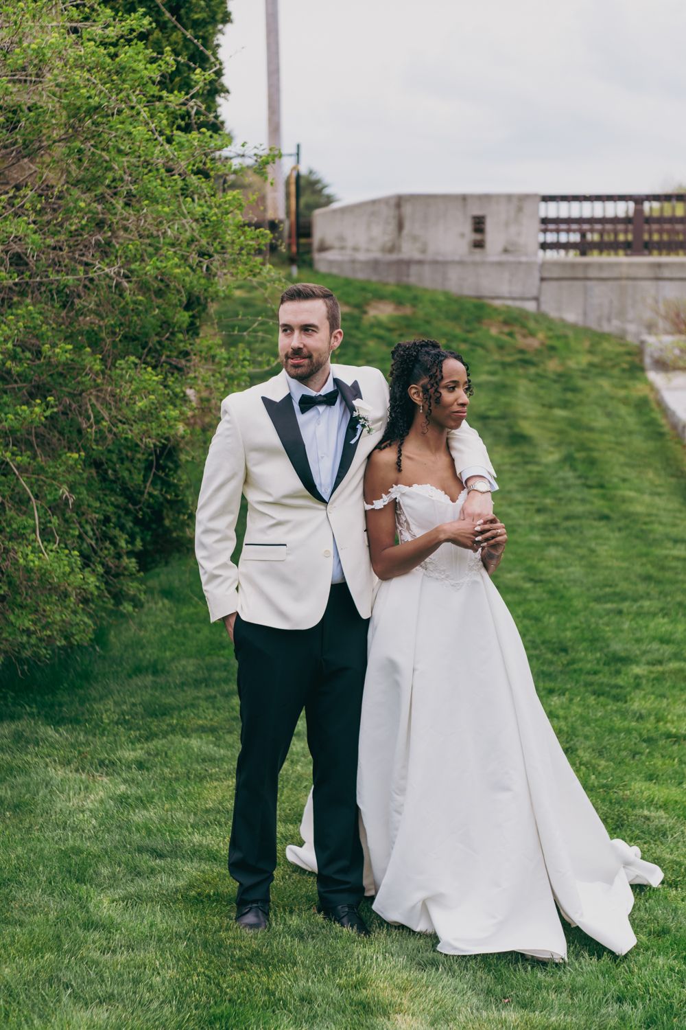 Interracial couple in wedding attire posing on green grass, groom in white tuxedo, bride in off-shoulder white gown.