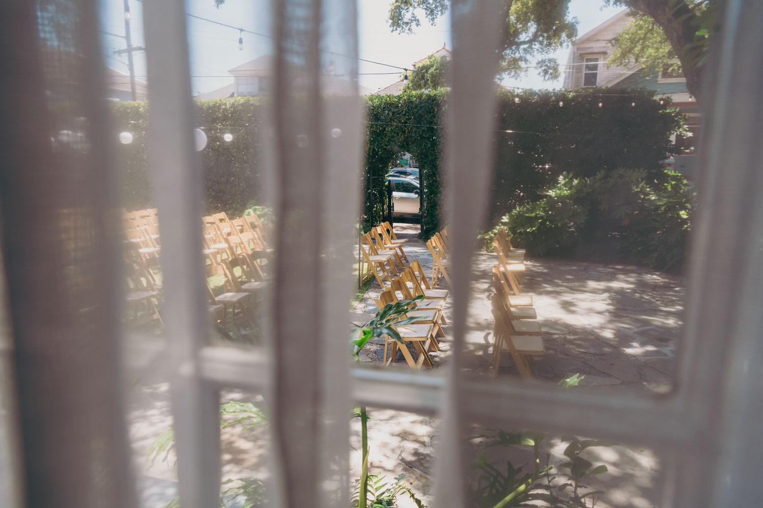 View through a sheer curtain of a sunlit garden patio with wooden chairs and lush greenery.