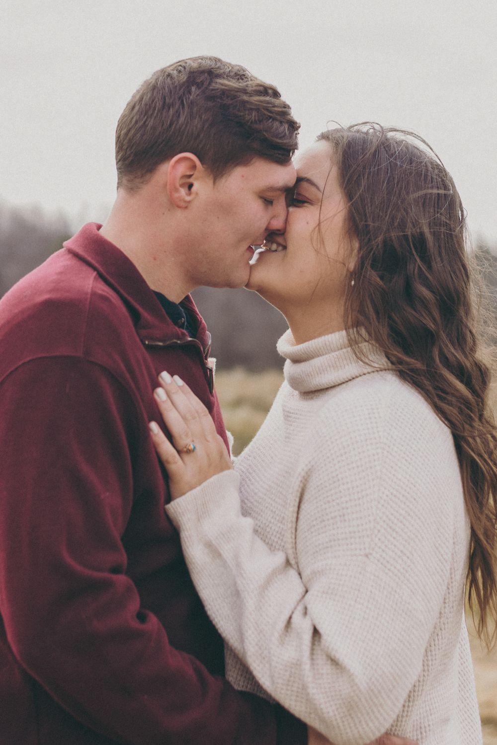 A couple sharing a romantic almost-kiss outdoors, woman in white sweater and man in burgundy pullover touching foreheads.