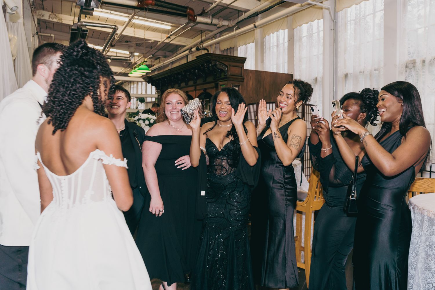 Bride surprises bridesmaids in black dresses who react with joy and applause at wedding reception.