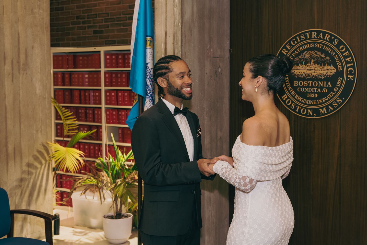 Couple exchanging vows at Boston City Hall Registrar's Office, groom in tuxedo and bride in white off-shoulder gown.