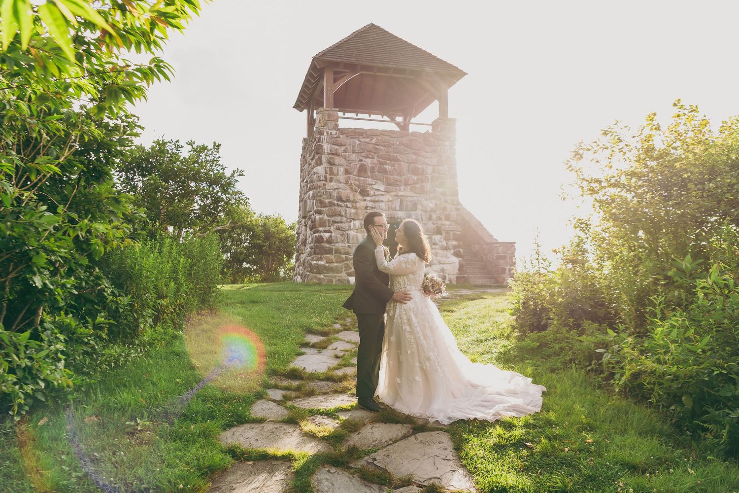 Bride and groom embrace on stone path beside rustic stone bell tower surrounded by lush greenery in golden sunlight.