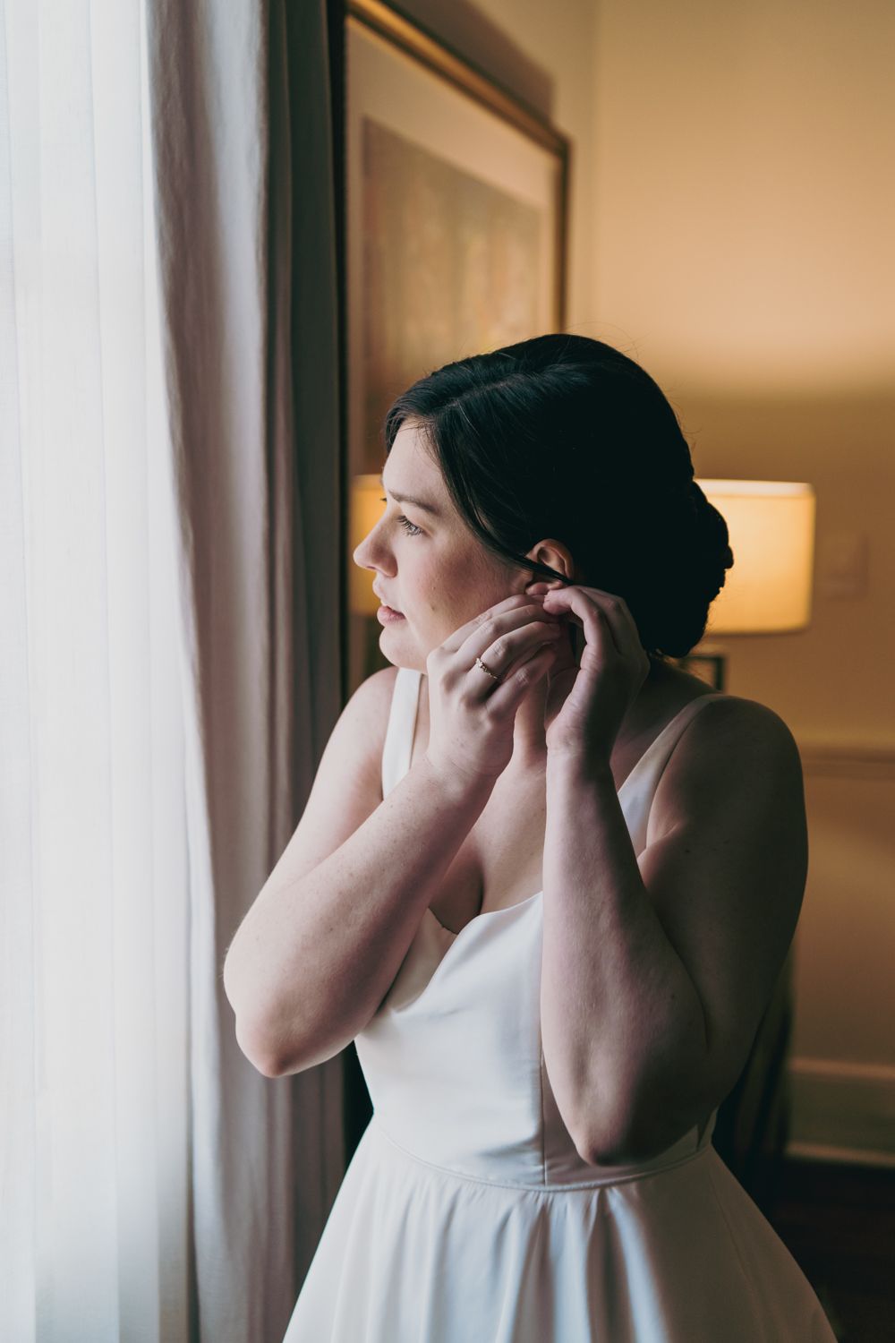 Bride in white gown putting on earrings by a window with soft natural light.