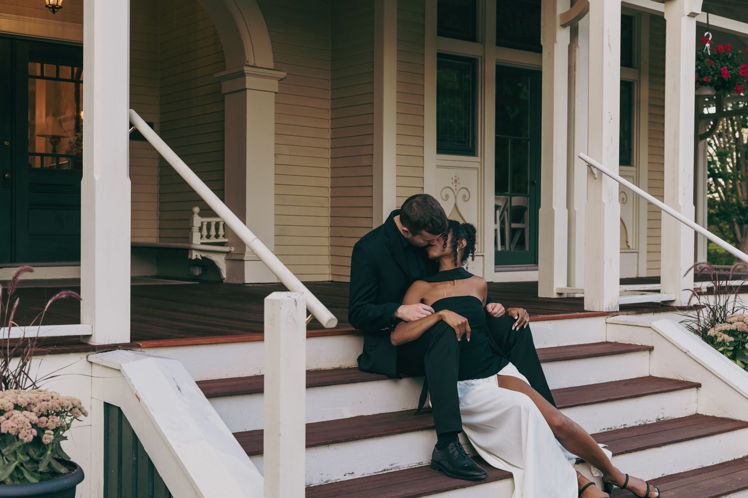 Couple in formal attire sharing a romantic kiss on white wooden porch steps of a charming Victorian home.