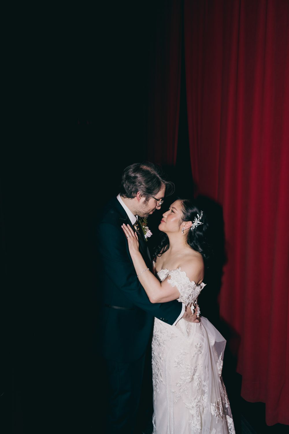 Bride and groom share an intimate moment in front of a dramatic red curtain backdrop.