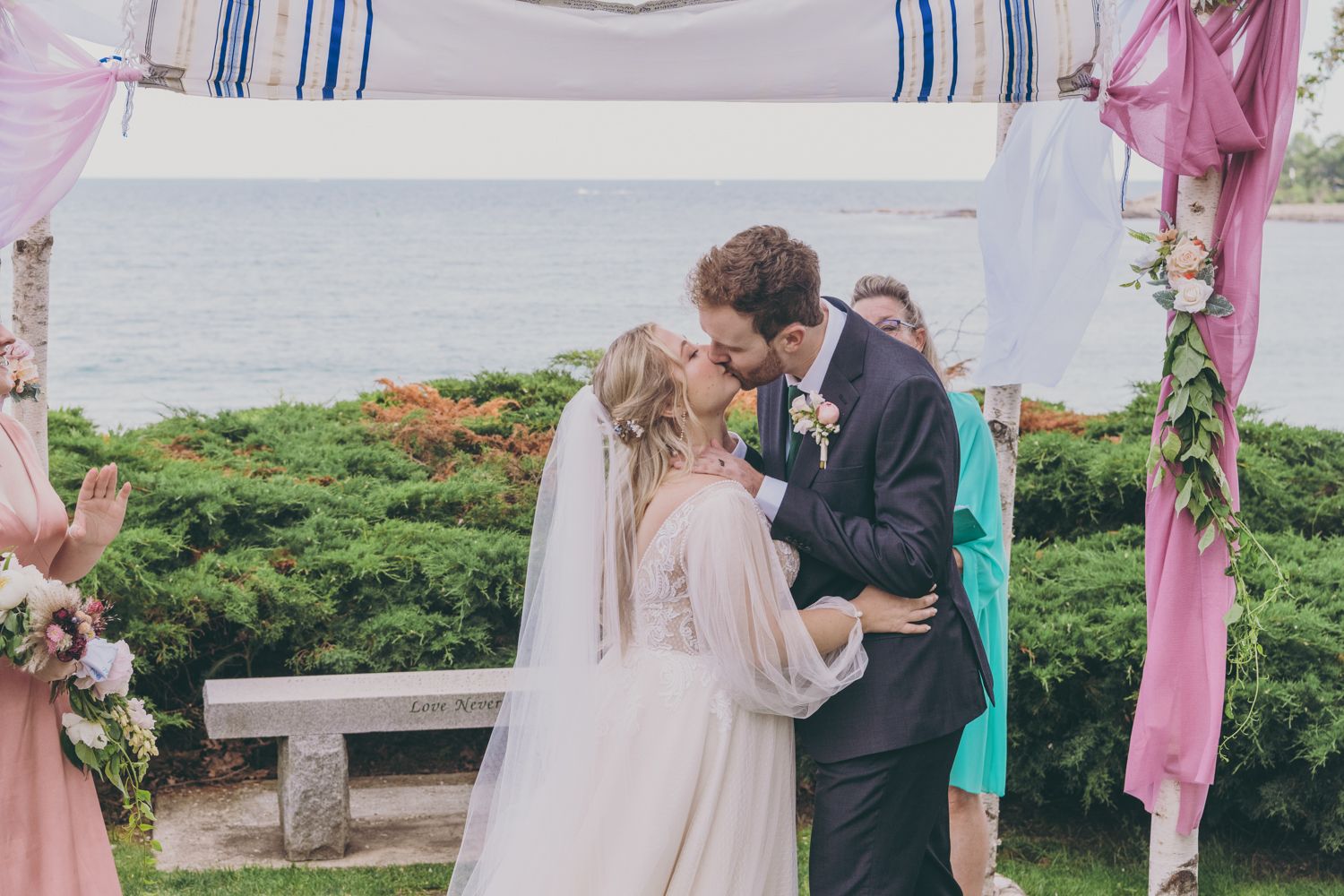 Bride and groom share their first kiss under a decorated wedding chuppah with ocean views behind them.
