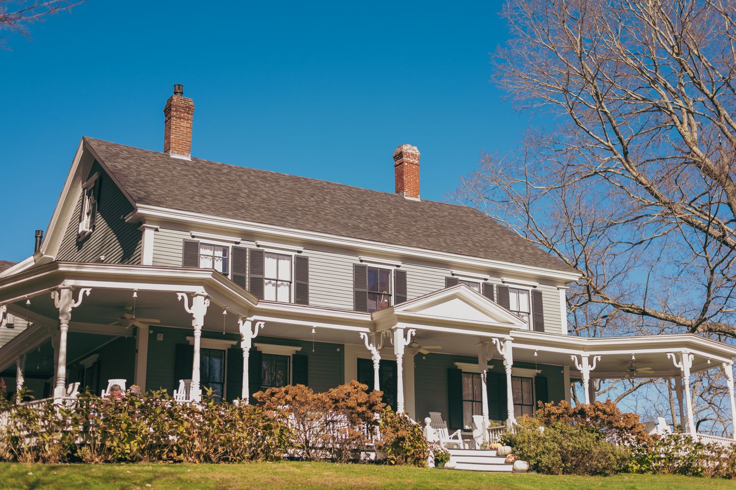 A charming gray Victorian farmhouse with wrap-around porch, shutters, and red brick chimneys under a clear blue sky.