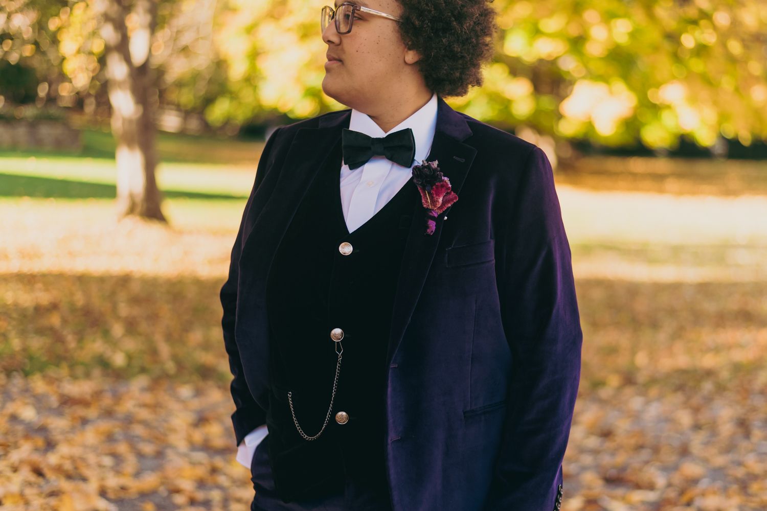 Dapper wedding spouse in purple velvet suit with bow tie and boutonniere among fall foliage.