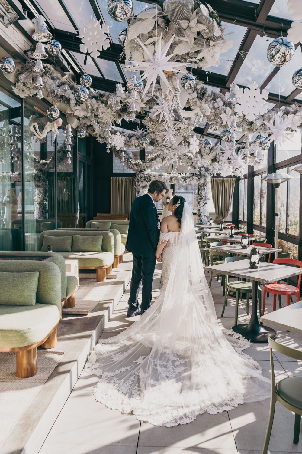 A bride and groom stand together in an elegant sunlit venue adorned with white floral ceiling installations and a long lace veil.