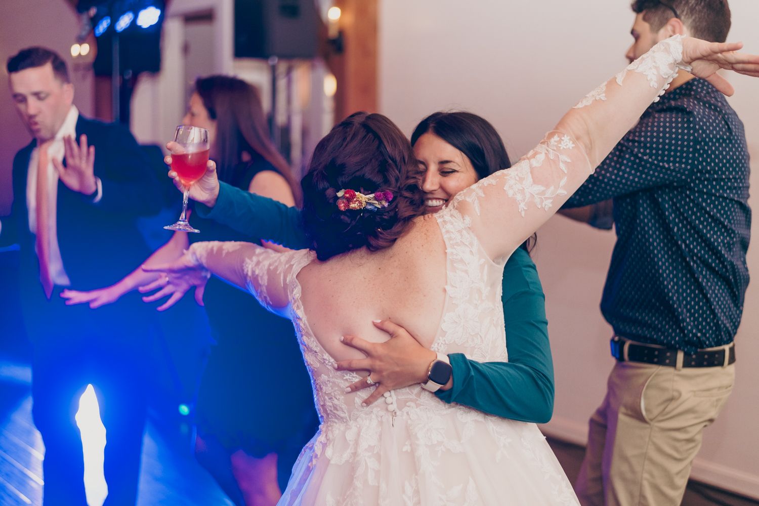 Bride in lace wedding gown shares joyful hug with guest on dance floor during wedding reception.