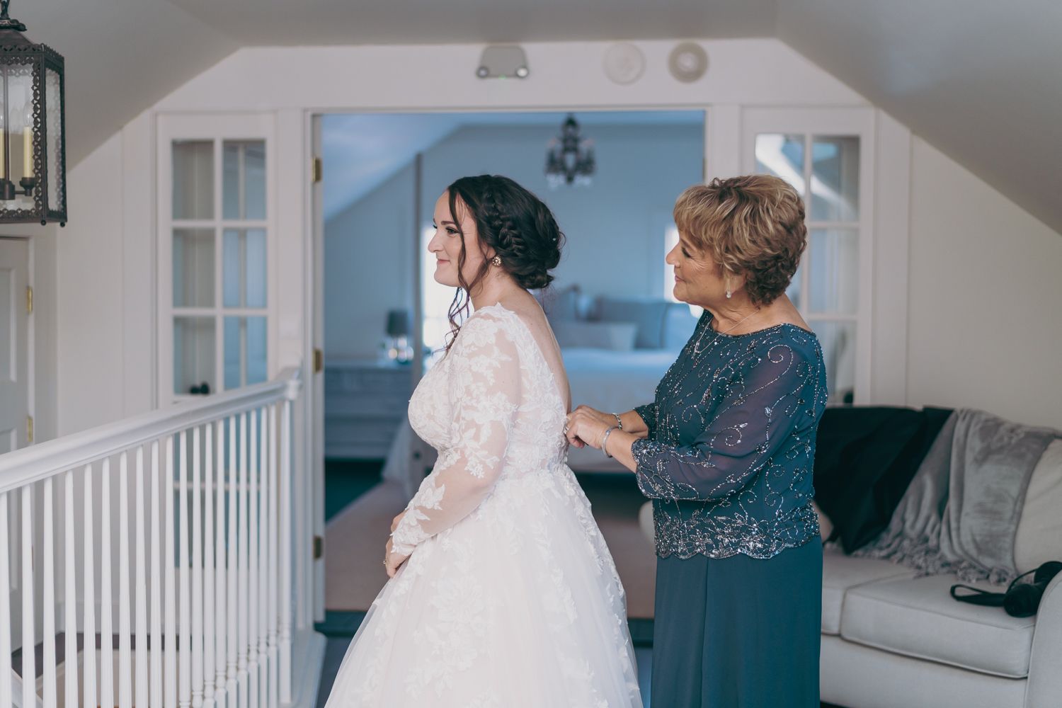 Bride in white lace gown getting dressed with help from older woman in blue dress indoors.