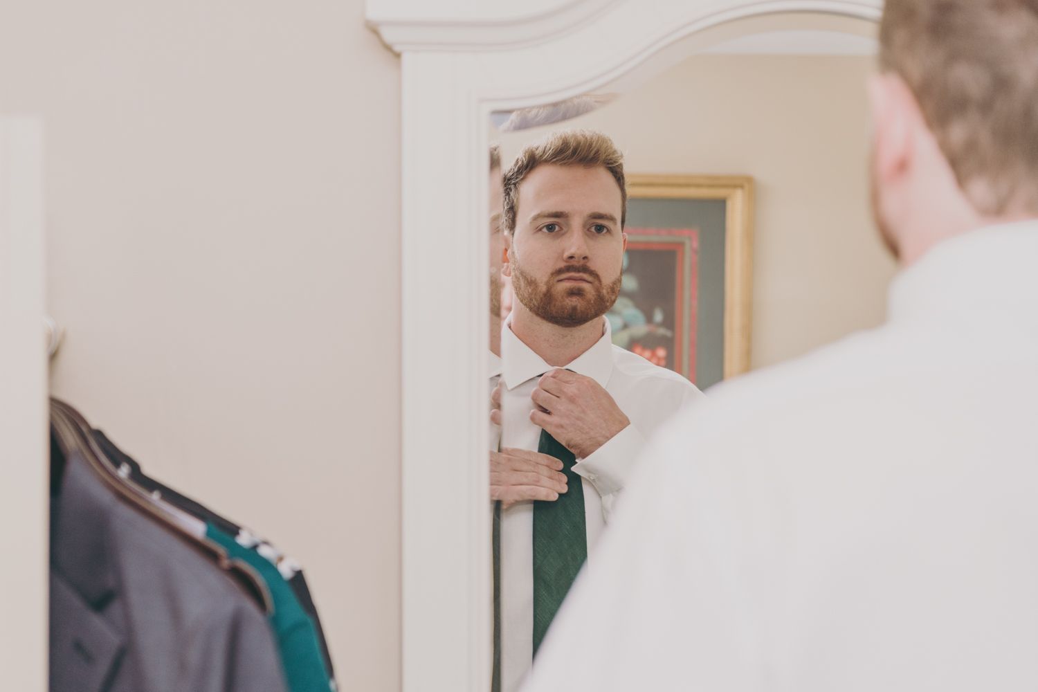 Groom adjusting his tie while looking at his reflection in a mirror while getting ready for wedding.