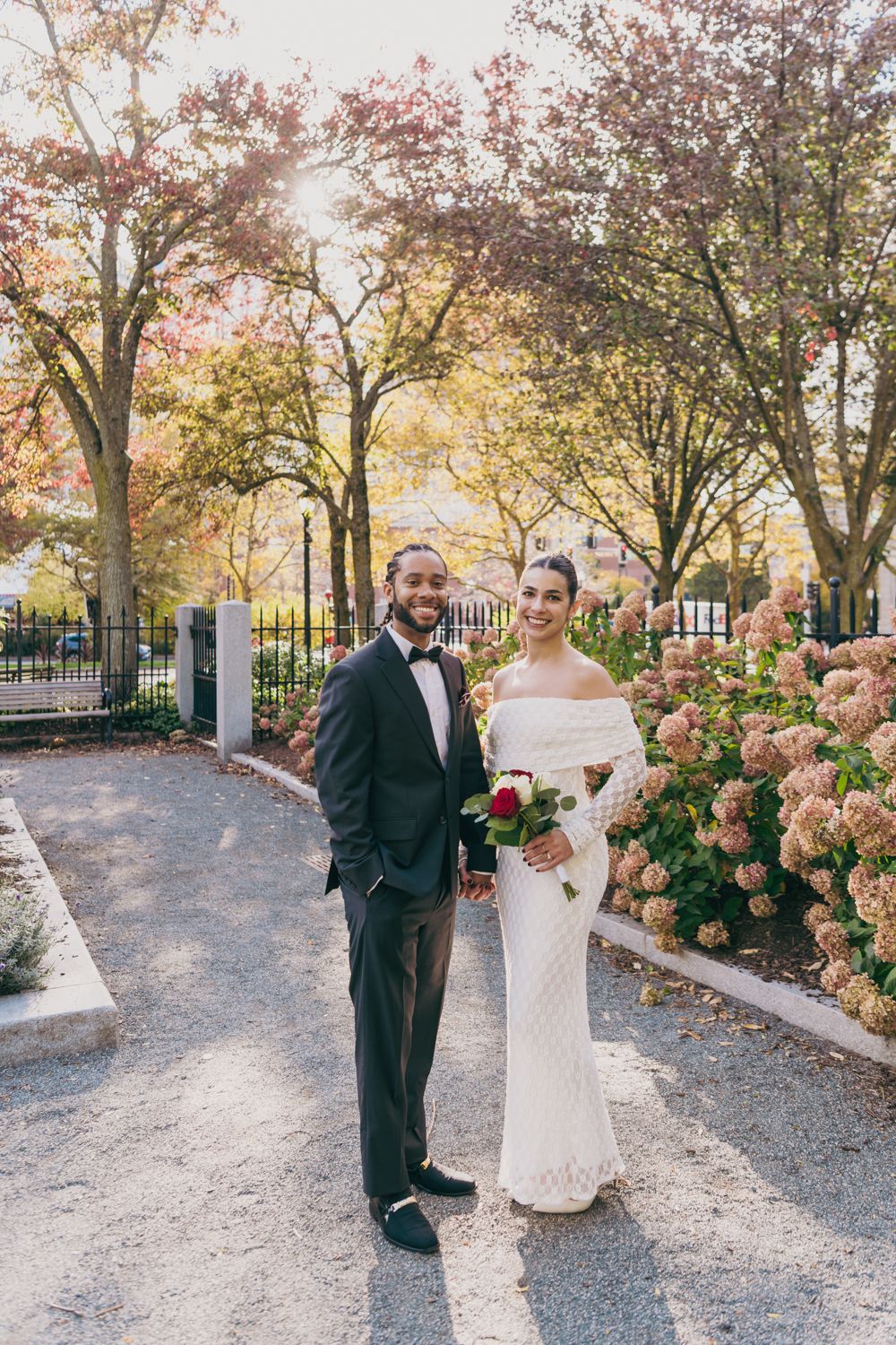 Couple in wedding attire posing in a blooming park, groom in tuxedo and bride in white off-shoulder gown holding red bouquet.
