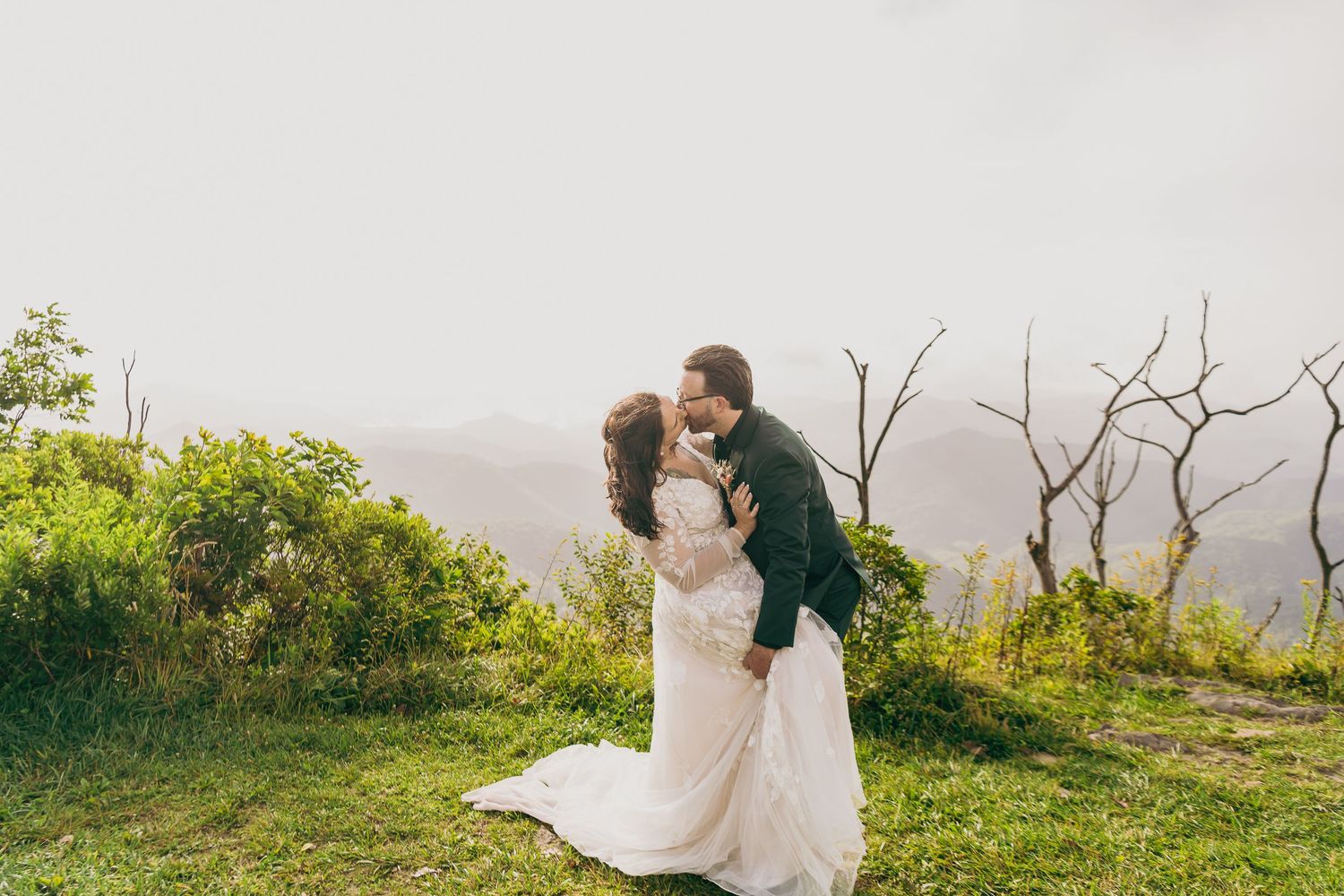 Bride and groom sharing a romantic kiss on a scenic mountain overlook, bride in elegant lace wedding gown.
