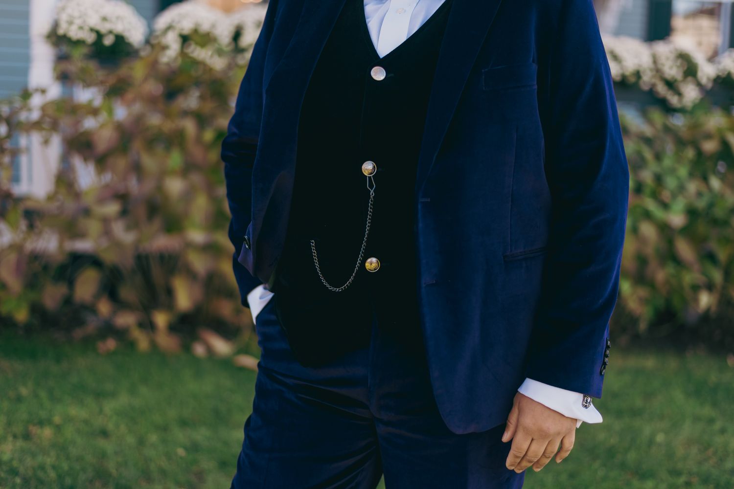 Groom in navy blue velvet suit with gold chain detail and boutonniere outdoors.