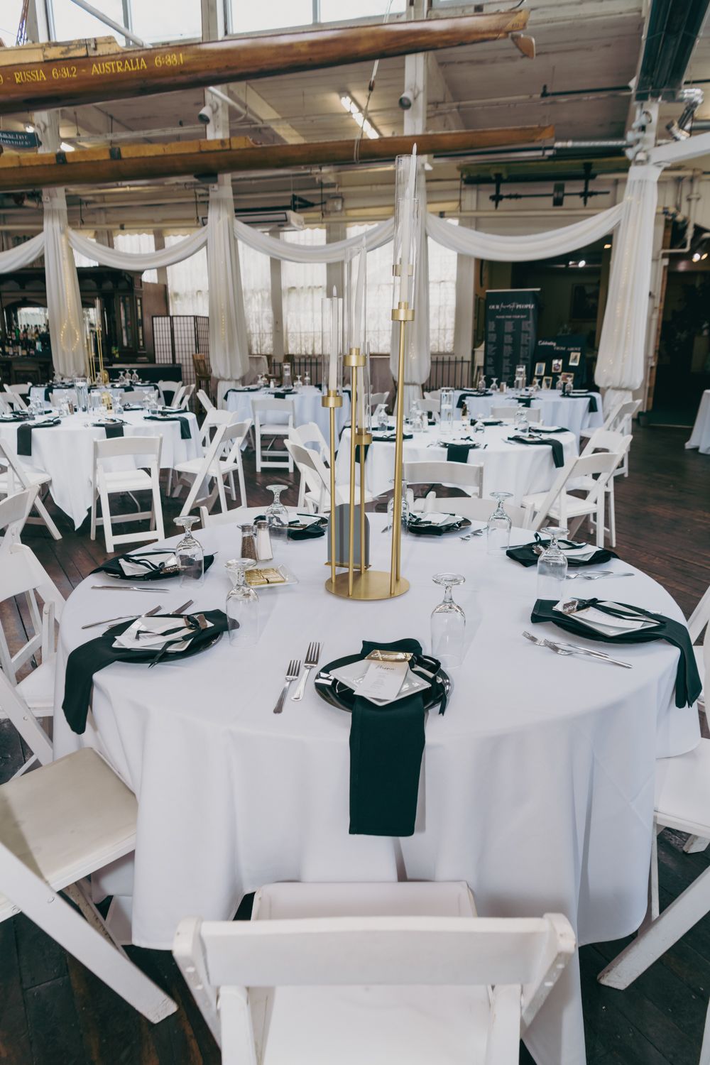 Elegant wedding reception tables with white linens, black napkins, gold candelabras, and white folding chairs in an industrial venue.