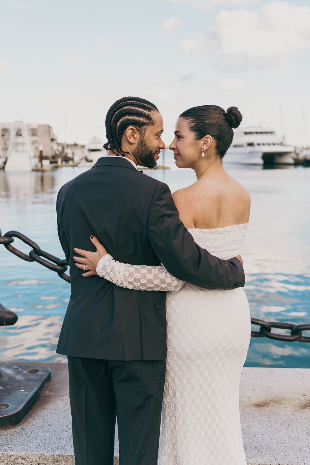 Newlywed couple embracing near marina waterfront, groom in black suit and bride in lace gown.