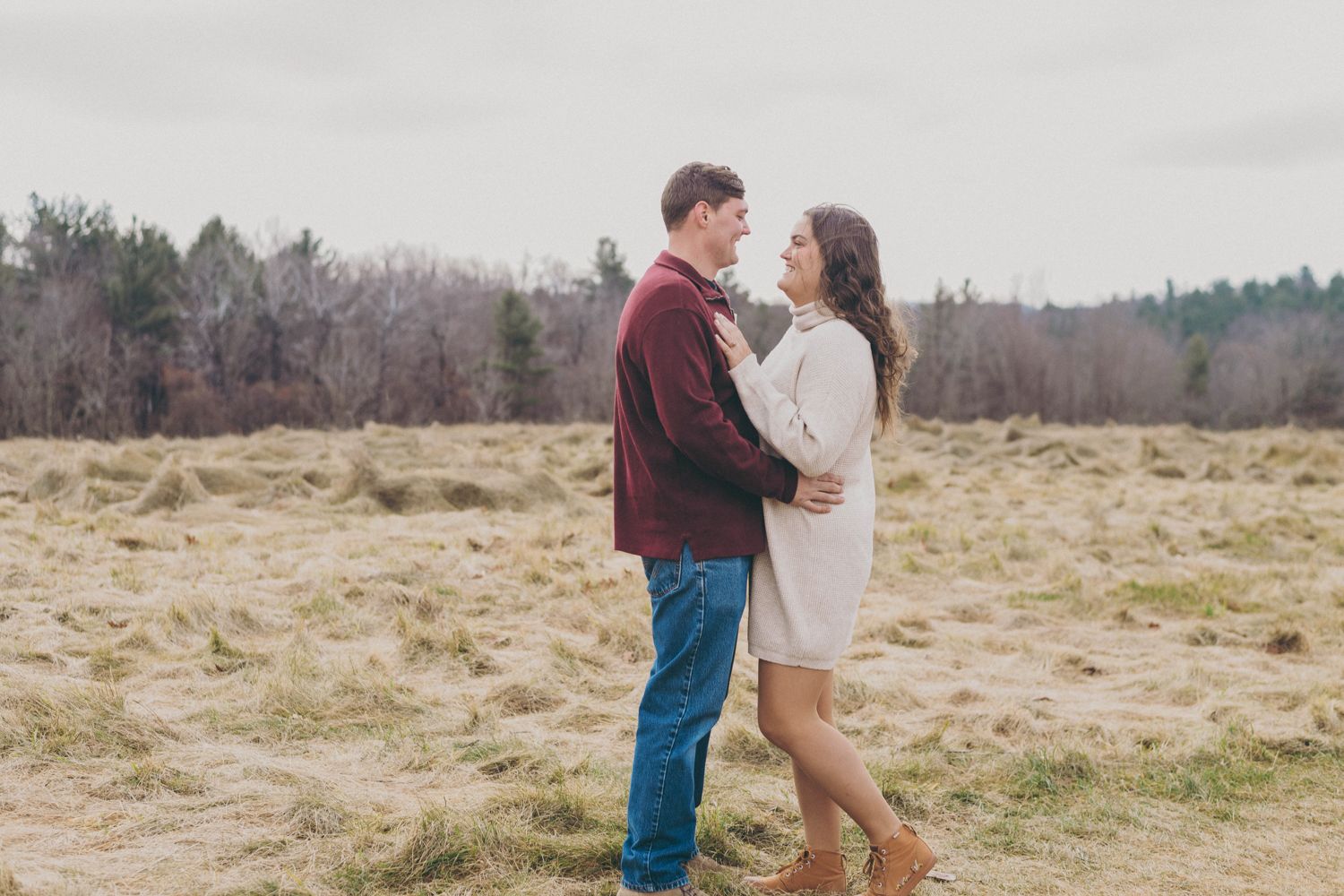 A couple embracing in an open field during autumn, man in burgundy shirt and jeans, woman in white dress.