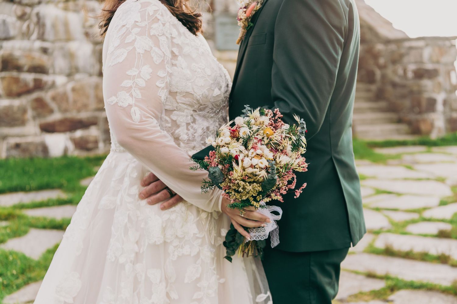 Bride in lace gown and groom in dark suit holding a wildflower bouquet at a rustic stone wedding venue.