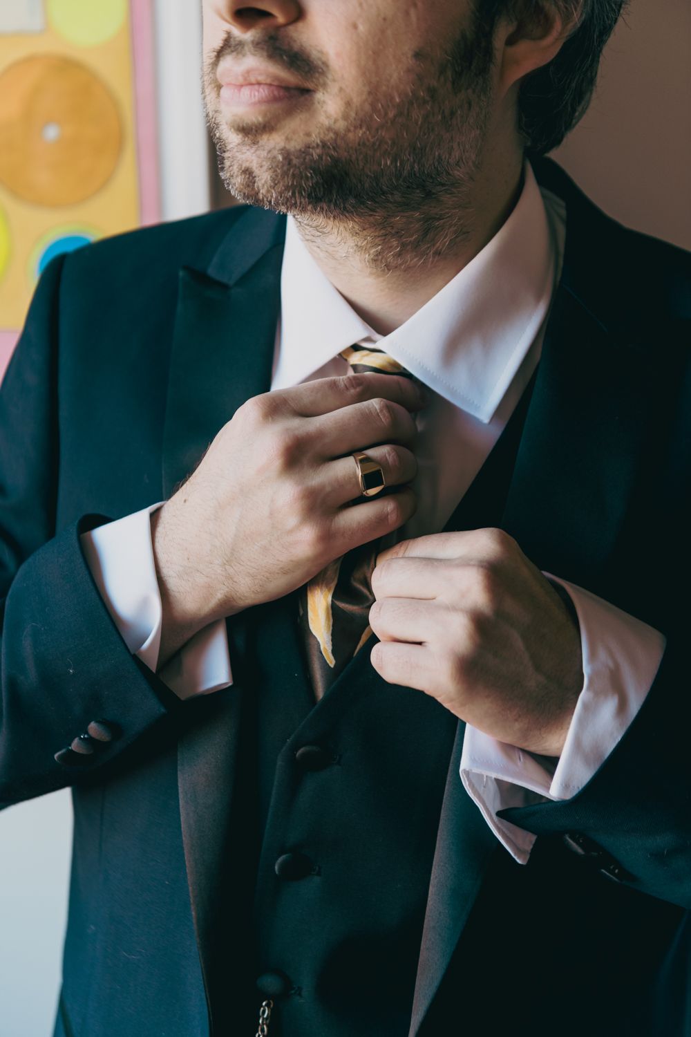 Man in dark suit adjusting gold striped tie, wearing signet ring, white dress shirt, formal wedding attire.