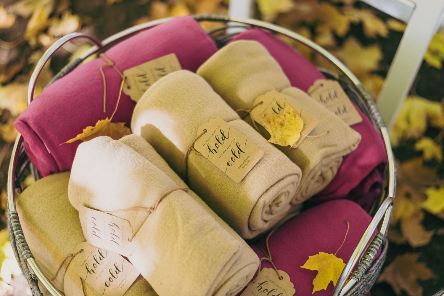 A basket of rolled blankets in pink, yellow, and cream colors with tags, displayed outdoors among autumn leaves.