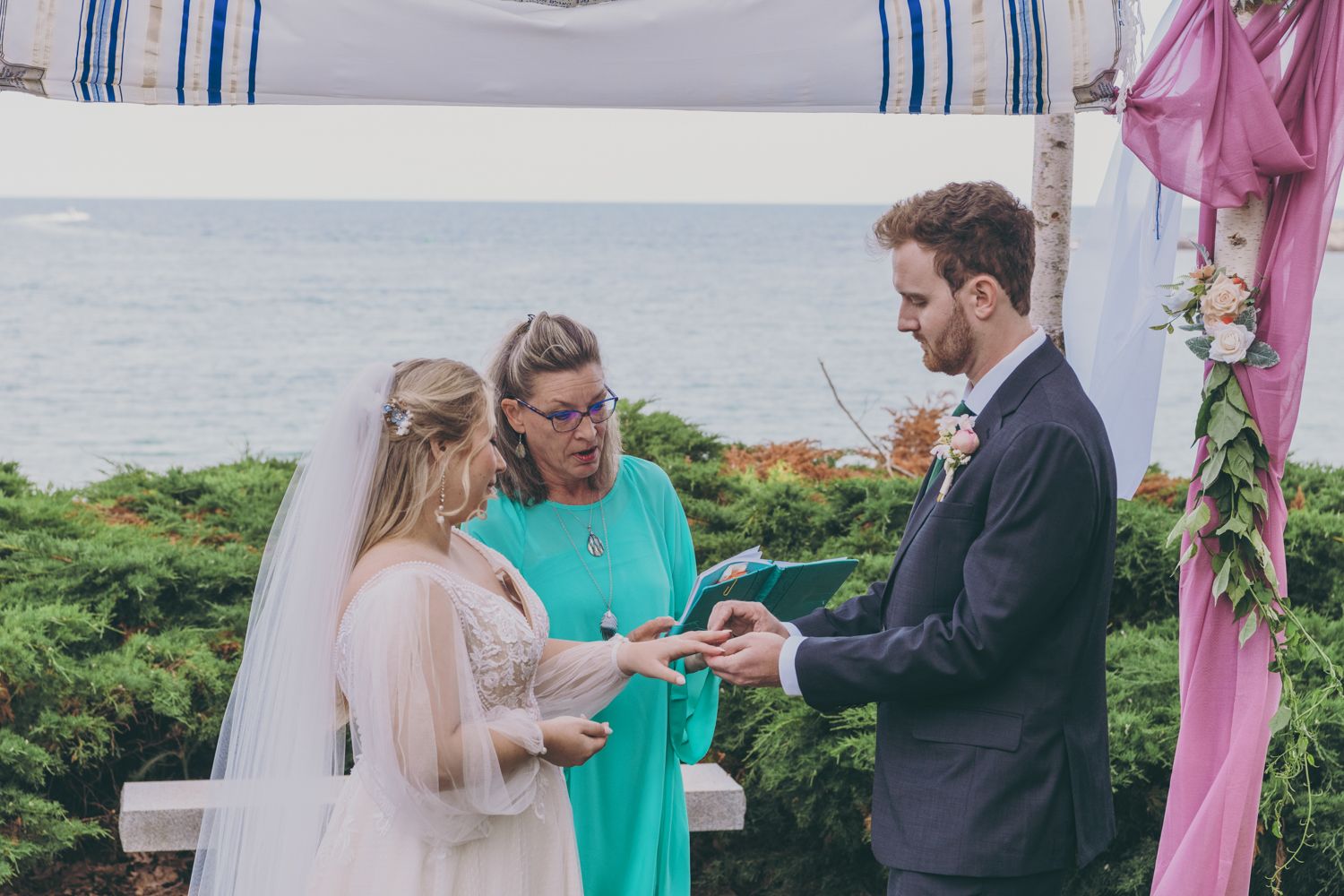 Bride and groom exchange rings during outdoor wedding ceremony by the ocean with officiant in teal dress.