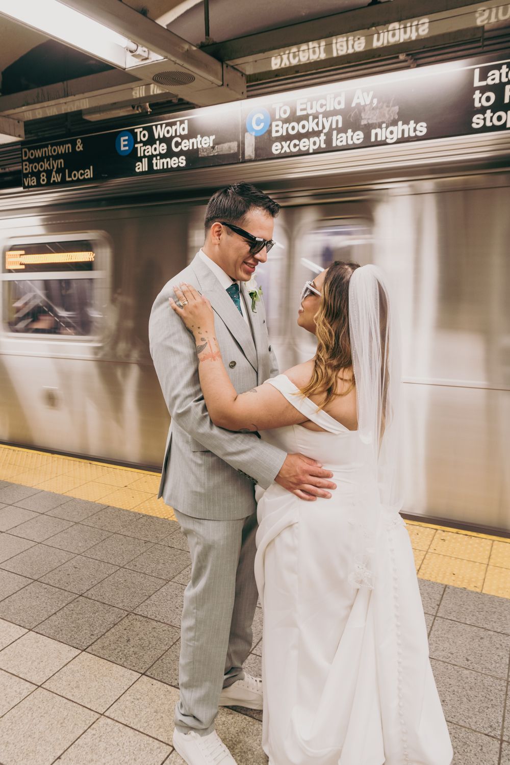 Newlywed couple embracing and smiling in NYC subway station, bride in white gown and veil, groom in gray suit.