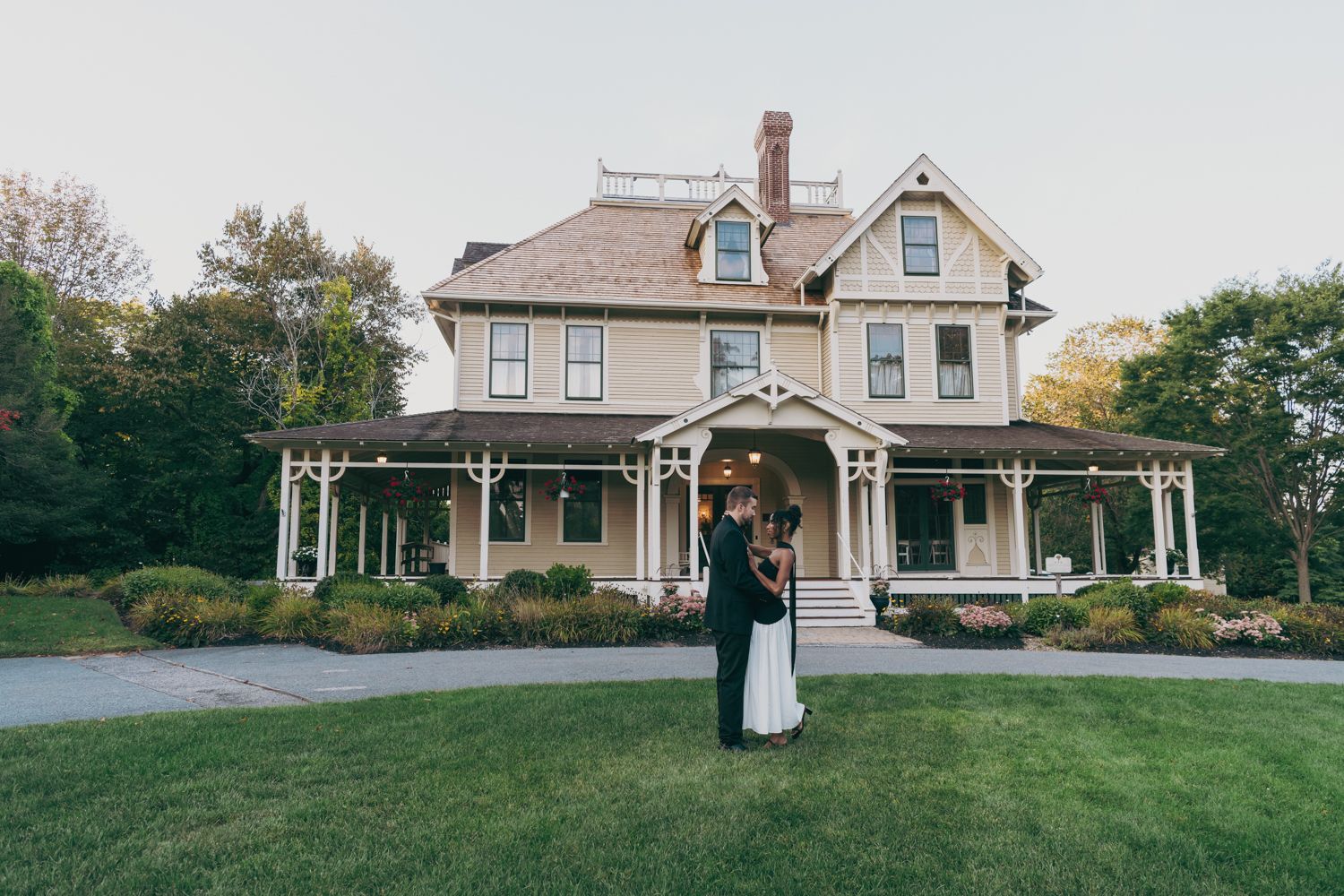 Interracial couple embracing on lawn before a large Victorian-style home with wrap-around porch at dusk.