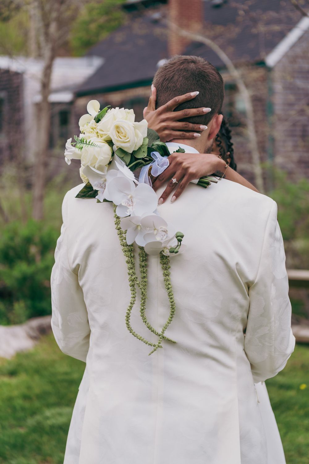 Bride holding white rose bouquet embracing groom in white suit from behind in garden setting.