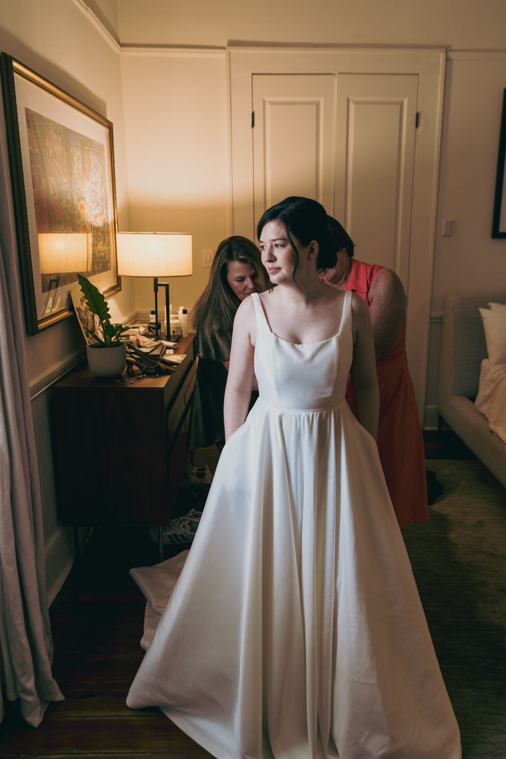 Bride in elegant white satin ball gown being helped by bridesmaids in a warmly lit room.
