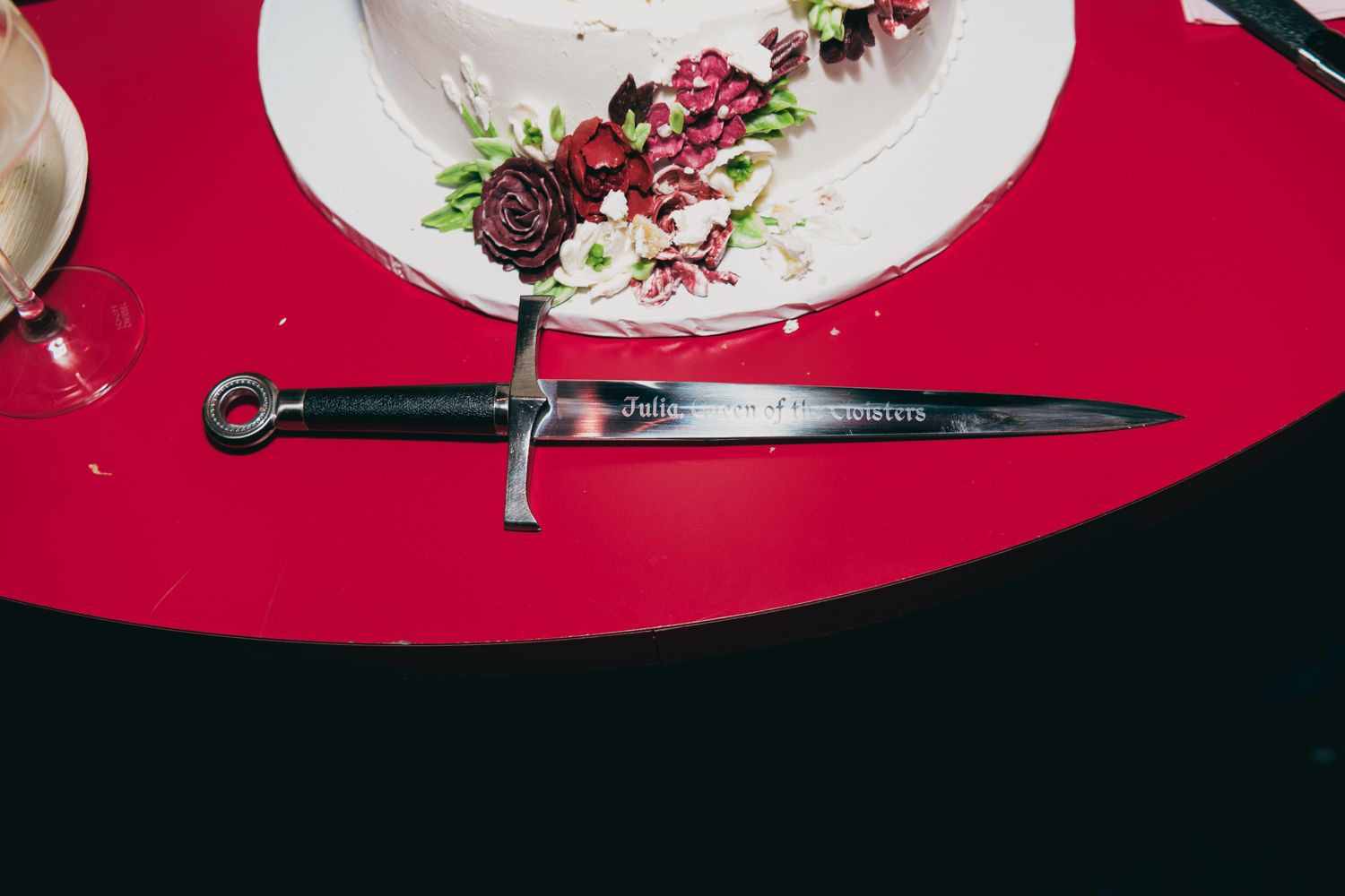 Ornate dagger cake cutting knife on red table beside floral wedding cake.